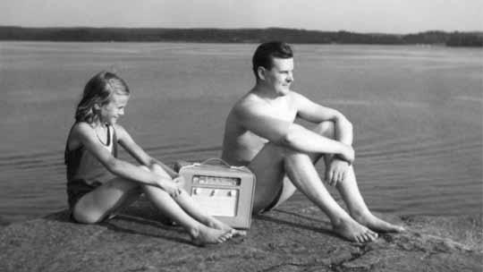 Isä ja tytär kuuntelevat matkaradiota veden äärellä. Kuva sarjasta Kun radio tul